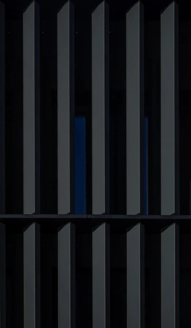 Close-up view of a modern building façade with vertical dark metal fins, creating a repeating geometric pattern with narrow gaps that reveal a deep blue background.