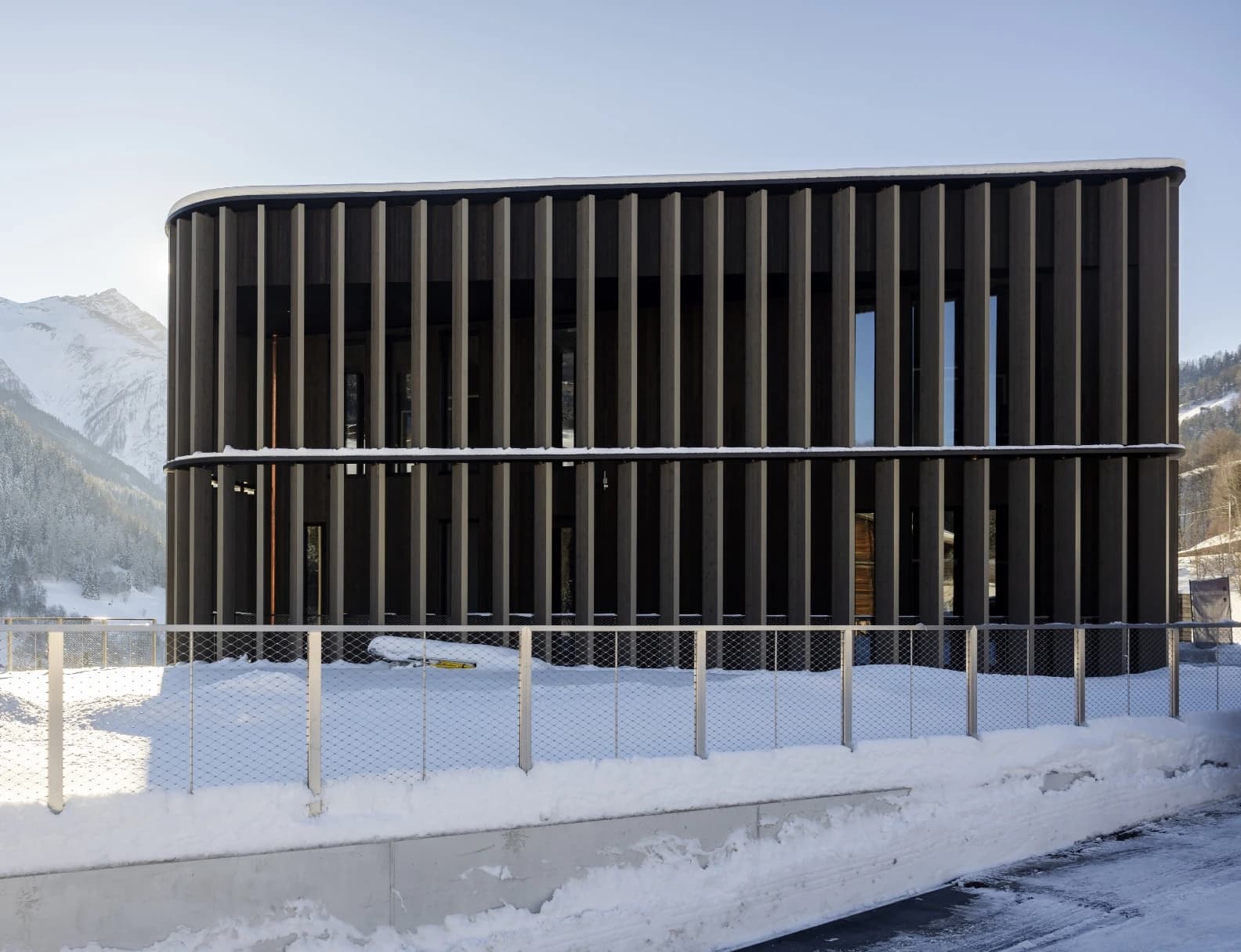 Modern two-storey building with dark vertical timber fins and rounded corners, snow on ledges, set against alpine mountains.