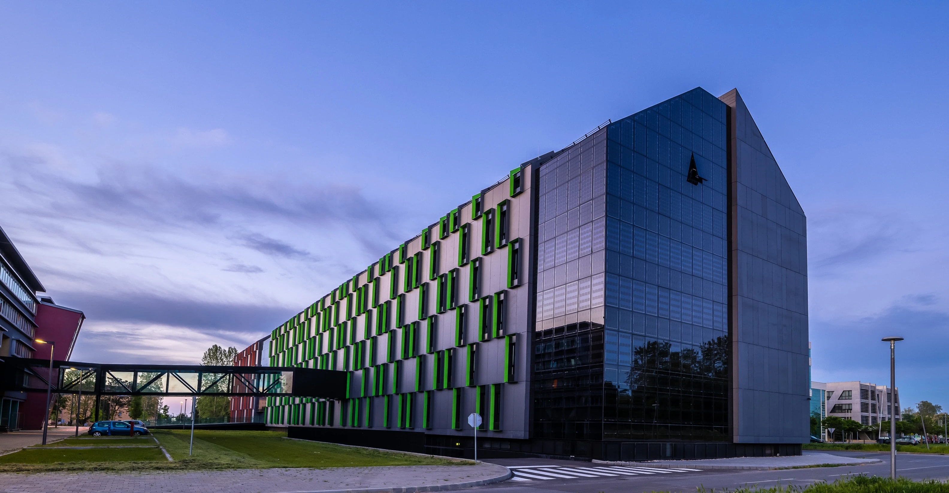 Modern university campus building with a sleek black glass facade and green-accented panels, connected to a neighboring structure by a glass walkway, photographed at dusk under a blue and purple sky.